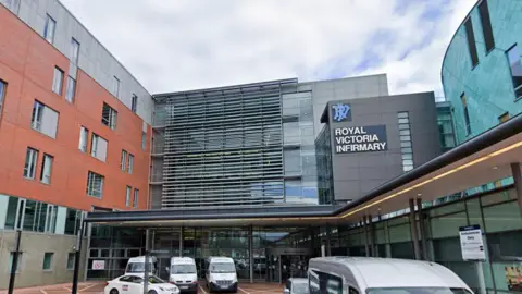 An exterior view of a hospital building with cars and ambulances outside and a sign saying Royal Victoria Infirmary