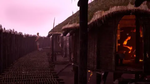 Guy Schofield A digital art installation shows people in wooden huts and one person standing on a pathway on the left. Inside one of the huts, which is closest and to the right, there is a glowing orange light from a fire.