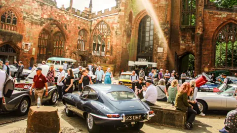 People enjoying looking at a display of vintage cars, including a Jaguar E-Type, in the Coventry cathedral ruins.