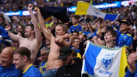 AFC Wimbledon fans dressed in blue and yellow, including two men who are topless, celebrate and punch the air in the stands at Wembley Stadium.