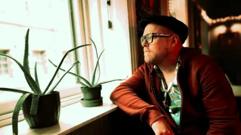 Phil Jackson sits next to a window and looks outside. He has a black cap on, glasses, a dark orange jacket and top underneath. There are two plant pots sitting on the windowsill next to him. 