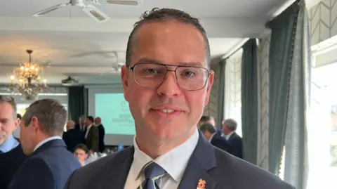 Marcus Calvani dressed in a dark suit jacket, white shirt, and striped tie stands in the foreground of a bright event room. Several people are mingling in the background near a presentation screen. 