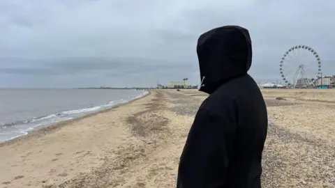 A picture of Leon in a black hoodie, with the hood covering his face to protect his identity. He is pictured on Great Yarmouth's central beach, with the Wellington Pier and big wheel visible in the distance.