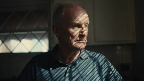 A man is looking through a window, with a long distant gaze.