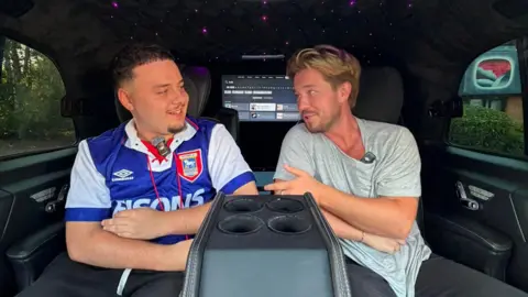 J2Hundred A man with a beard wearing an Ipswich Town football shirt sits in a car with another man wearing a grey t-shirt