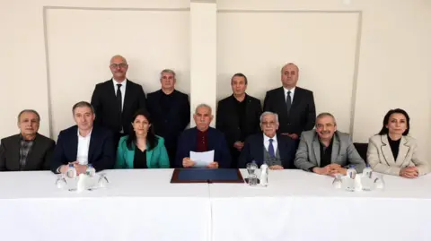 Dem Party Abdullah Ocalan, holding a piece of white paper in front of him, sits in the centre of a group of pro-Kurdish MPs, with a table in front of them covered in a white tablecloth