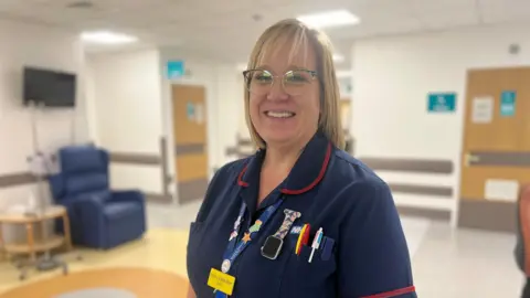 NHS Somerset Foundation Trust Emma Wells-Burrs is looking at the camera. She's in a hospital ward, and is wearing blue NHS scrubs and glasses. She has colourful pens in her pocket and a blue lanyard on.