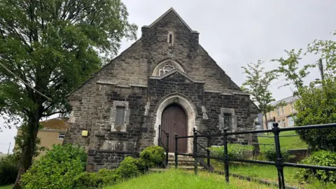Outside of Markham Congregational Chapel. A small grey stone chapel, on the side of a hill. A large arched wooden door is at the middle of the chapel, with two narrow windows either side. It sits among some grass and a path with railings. Trees can be seen either side of the chapel, with a row of houses up the hill behind it.
