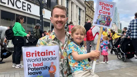 Nicola Rees/BBC A man wearing a hawaiian shirt kneels with his young son on his lap. They are holding placards saying "Britain: world leader in bad paternity leave" and "share the care". Both are smiling at the camera.