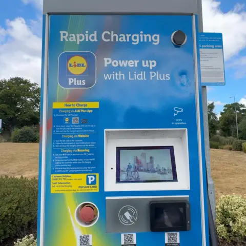 A blue Lidl Plus rapid electric vehicle charging unit stands in a car park at Lidl's Barton-upon-Humber store. The charger displays instructions on how to charge using the Lidl Plus app, website or roaming services. A controlled parking area sign is mounted on a post behind the charger, while grass, shrubs and trees can be seen in the background beneath a partly sunny sky.