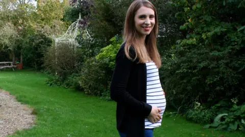 Ali Harris Ali Harris is standing outdoors in a peaceful garden setting. She’s wearing a black cardigan over a white and blue striped top, paired with blue jeans. Her long, straight hair frames her face, and she’s smiling at the camera. One hand rests gently on her stomach, she is pregnant. Behind her, the garden is full of greenery, there are trees, bushes, and a grassy lawn. A white, decorative metal structure and a gravel path are also in the background.