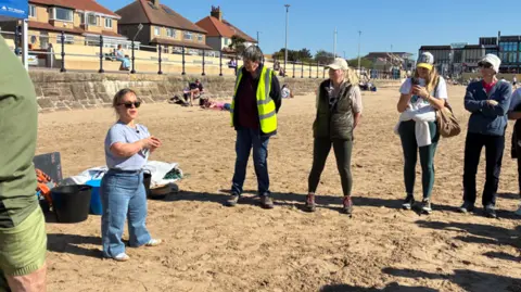 Ellie Simmonds who is wearing jeans and a t-shirt and sunglasses talks to volunteers ahead of a beach clean on West Kirby beach on a sunny day.
