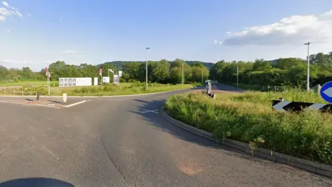 Google A Google Streetview image of a roundabout. A grassy area and trees are on the other side of the road.