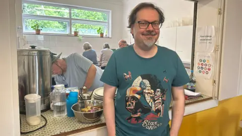 Cad Taylor, BBC Man stands in front of a kitchen hatch, he has glasses and wears a tshirt