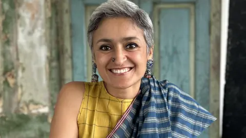 Chetna Makan A woman smiling and looking at the camera. She is wearing large silver earrings and has a silver nose piercing. She is wearing a yellow top with a blue and white shawl draped over it. In the background, which is out of focus, there is a blue door.