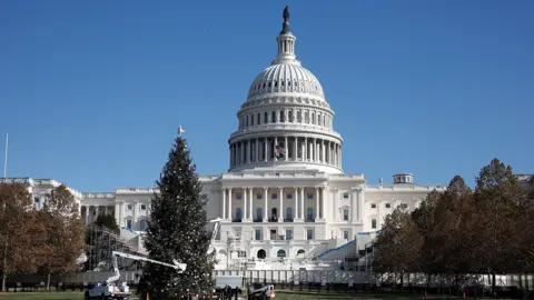 A wide, front-on view of the US Capitol against a blue sky, with a Christmas tree being installed in front of the building, taken in Washington, DC on 2 December