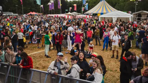 Menter Caerdydd Swarms of people on a field at Tafwyl, with tents in the background and a barrier in the foreground with one group of young people waving at the camera