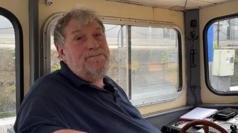 Dan Bater/BBC Simon Tipple is seated at the steering wheel of a boat. He has grey hair and a short beard and is wearing a dark polo shirt.
