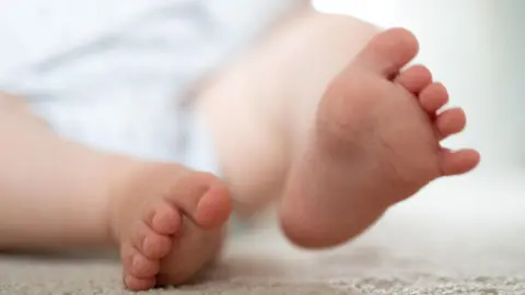 A baby's feet - one on a cream coloured mat or blanket and the other lifted slightly in the air.