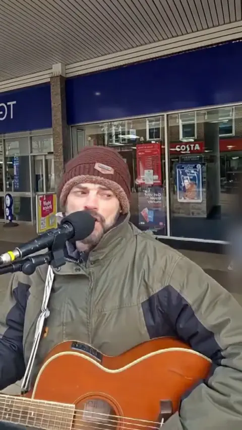 Wes Dolan, a busker, playing an acoustic guitar outside a shop. He is wearing a green jacket and a brown hat.