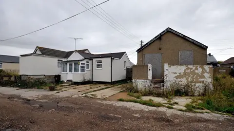 Cracked pavements and roads in Jaywick. In the background are bungalows with peeling paint and one has boarded up windows.