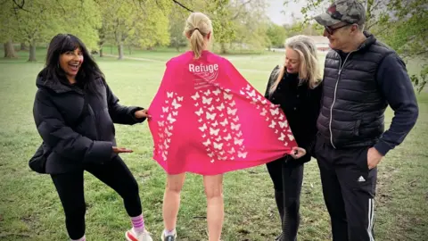 Hetti Barkworth-Nanton Hetti Barkworth-Nanton is standing with her back to the camera between three people who are all dressed in black. She is showing the camera the butterflies on her pink cape while her friends point to it and smile at the camera. They are all standing in a field.