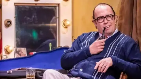 Pamela Raith Photography Bob Golding as Eric Morecambe reclining in a chair and half smiling with a prop pipe in his mouth. He is wearing a navy blue cardigan with some white stripes on it. There is a dressing room mirror behind him as well as an open violin case.