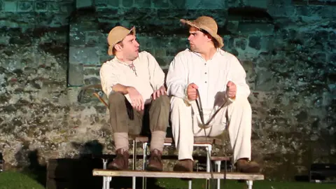 Packing Shed Theatre Two male actors performing while perched on what looks like a bench in front of a brick wall belonging to a castle. They are looking at each other.