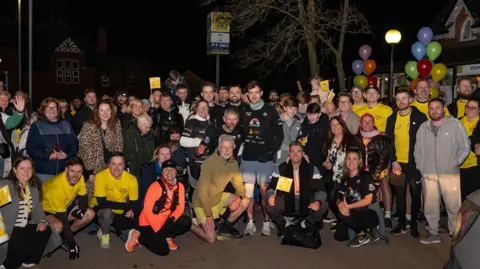 Merseyrail/PA Wire Alex Rigby, pictured in a black hoodie and surrounded with supporters after reaching Hunts Cross station - the final stop of his running challenge. The crowd have flags and balloons.