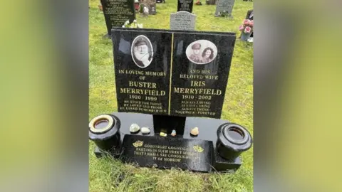 Verwood Town Council A black grave stone dedicated to husband and wife Buster and Iris Merryfield. A small model of a yellow robin reliant and figure of Uncle Albert sit on the base of the headstone
