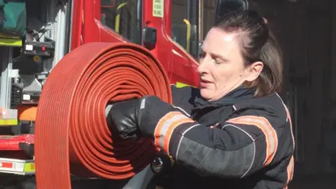 Ms Bourne is standing outside of a fire truck in her full fire black fire uniform. She has medium length brown hair. She is holding a large red fire hose and rolling it up around itself. 