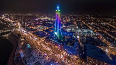 The Blackpool Tower is illuminated in pink, yellow, green and blue, including a heart near to the bottom of the tower. Lights and crowds of people and cars can be seen along the promenade area and on the road in this aerial photo.