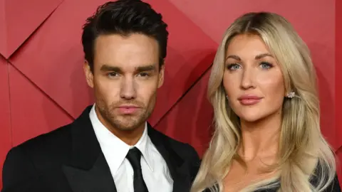 Liam Payne in suit and tie stands beside a smiling Kate Cassidy, with blonde hair and glittery earrings, against a red background