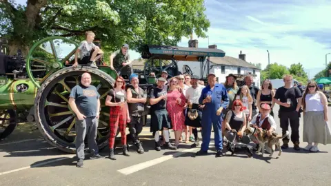 BBC A green traction engine with red wheels, eighteen people, two children and two dogs stand outside a pub. 