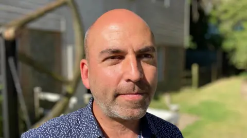 Luke Deal/BBC Tim Weller smiles at the camera. He is bald and has a shaved beard. He wears a blue shirt with some white details on it.