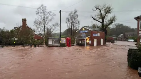 A village that is flooded. Dark orange water covers the road and has submerged parts of a bus stop and telephone box. The water is covering the entrance to a fish and chip shop. 