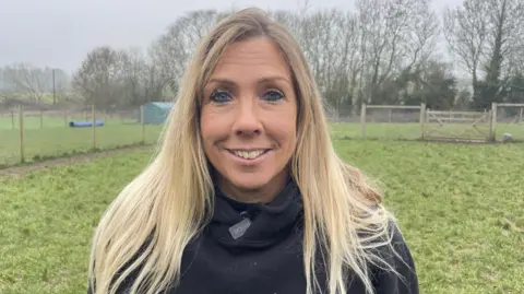 A woman with long blond hair smiles at the camera as she stands in a paddock. She is wearing a black hoodie. She is standing in front of a grassy area with fences, which is used as her dog training paddock. There are trees in the background and the sky is grey and misty.