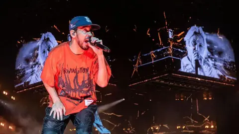 Getty Images Mike Shinoda's wearing a baggy red t-shirt and he's singing into a microphone with a cap. Behind him is a large screen with the band's singer Emily Armstrong.