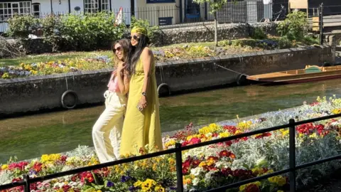 BBC/JO BURN Two women, one wearing a cream top and trouser set, the other a yellow sun dress, pose for a selfie in the bright flower beds of Westgate Gardens, just in front of the River Stour, with a moored punt behind them.