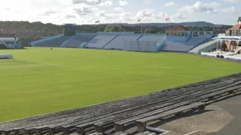 A large green cricket field with a stand of seats in the background, featuring a building with windows used for commentary and a scoreboard at the other end of the stand.