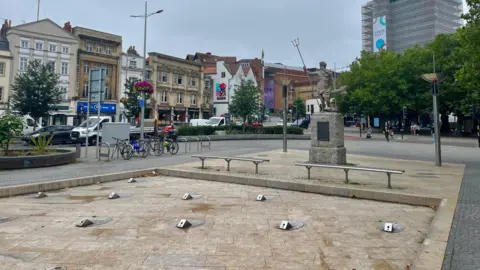 BBC Bristol City Centre fountains. It is a large, square, slightly sunken structure with small silver fountain nibs embedded in the stone and benches around the outside. There is no water flowing in the fountain.