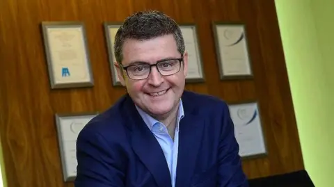 Brendan Mooney smiles while sitting at a desk with a Kainos sign above his head. He has short dark hair and black-framed glasses and is wearing a navy suit jacket and open-necked shirt 