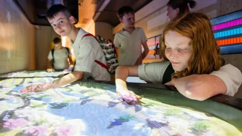 A group of schoolchildren try out an interactive exhibition in a railway carriage converted into a museum. The exhibit is a large sandpit on to which a railway line and landscape is being projected. In the foreground, a girl with ginger hair, wearing a white shirt, places her hand on to the sand. To the left, a boy with short dark hair, also wearing a white shirt with a backpack, places his hand on the sand. Other children can be seen in the background.