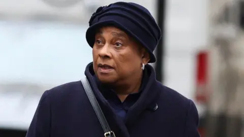 Baroness Doreen Lawrence dressed in a dark blue coat and hat. She has a black leather bag strap over her shoulder and silver earrings