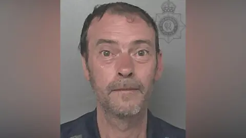 A police head and shoulders photo of a man with short brown hair and grey stubble looking directly into the camera. He is wearing a black top and the background is grey.