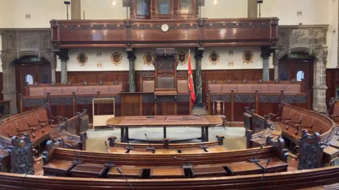 LDRS The Wakefield Council chamber has rows of dark oak seats, a table in the middle and a flag on a pole.