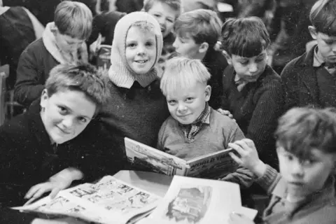 Getty Images A black-and-white photograph from the late 1960s showing a group of nine boys attending informal school lessons in Aberfan. The boys in the foreground are holding comic books, while one of the group has a light-coloured balaclava on his head. All of the boys are wearing school jumpers, and three of the group are smiling at the camera. 