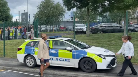 Outside shot of the school where it took place. A parked police car is seen , in front of the school gates. Two people walk past.