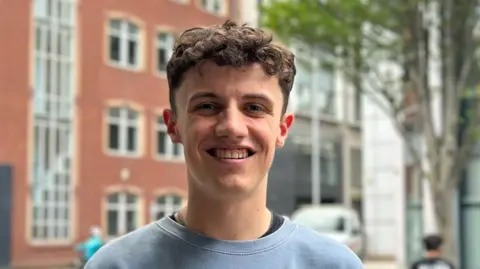 A young man with short curly brown hair smiles at the camera as he wears a blue sweatshirt with a black top beneath it. The background is blurred.