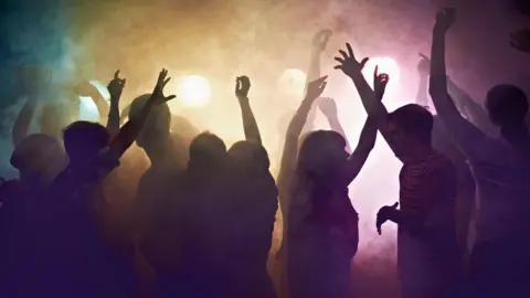Getty Images A crowd of people in shadow, dancing in multicoloured smoke in a nightclub with their arms in the air.